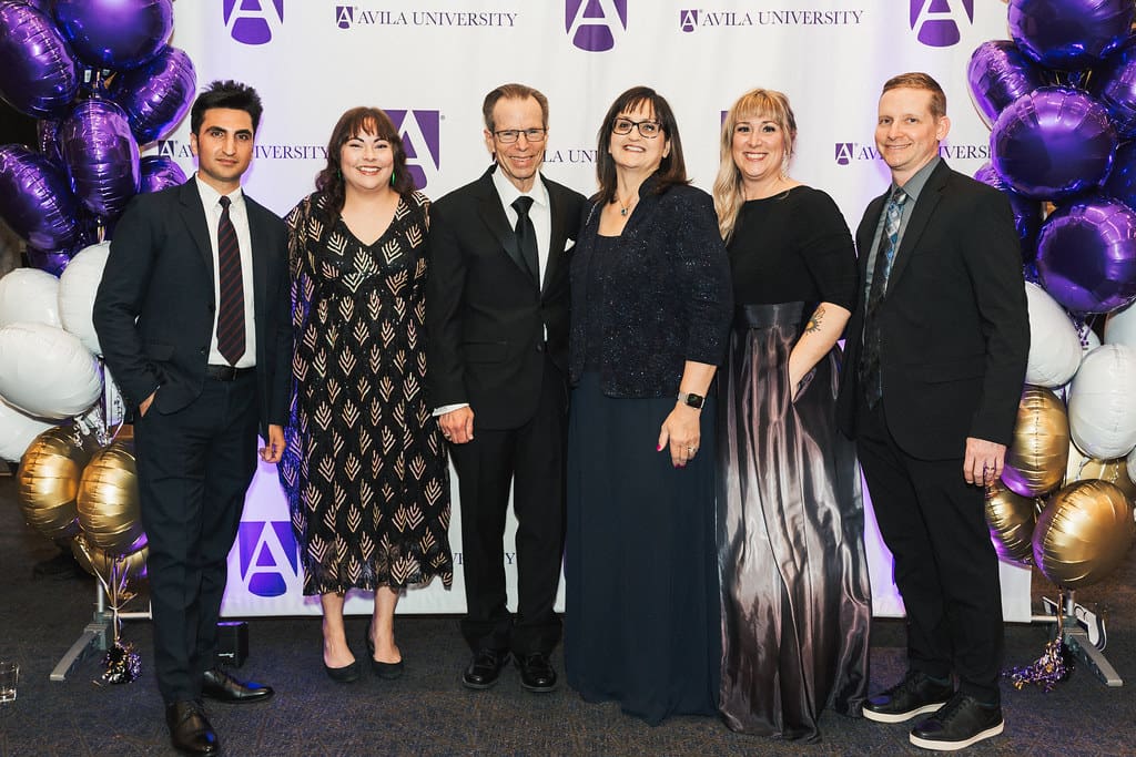 Six formal event attendees posing together in front of an Avila University backdrop.