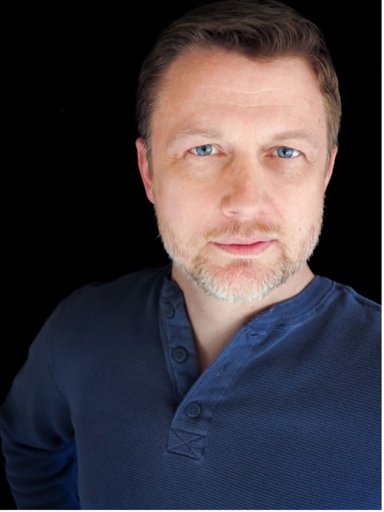Photograph of a person wearing a dark blue collared shirt against a black background. The person's face is obscured by a large blur effect, making facial features unrecognizable.