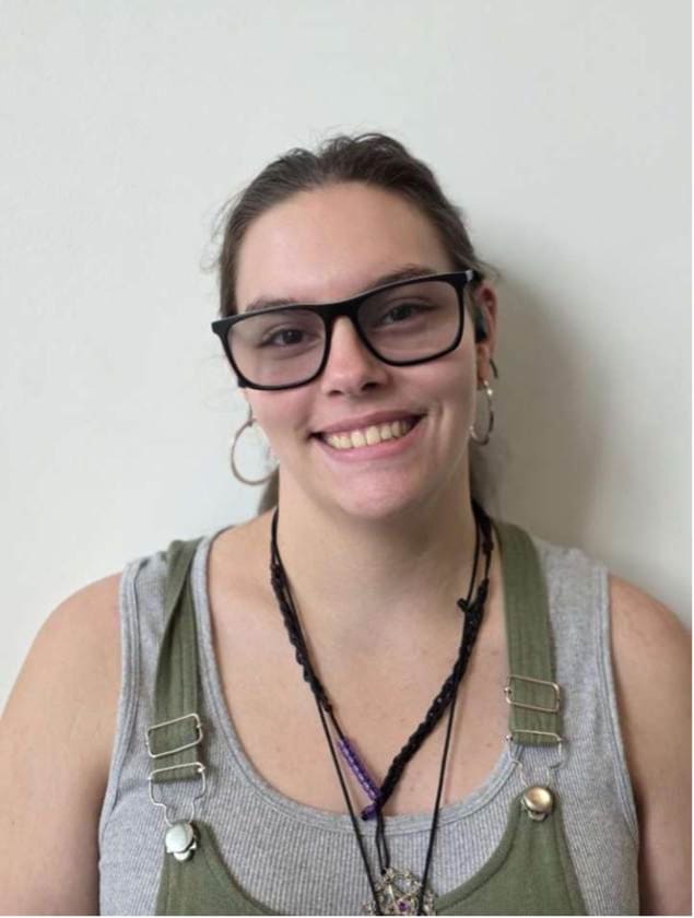 Photograph of a person wearing a gray tank top under green overalls with metal clasps and multiple necklaces. The person has earrings and glasses, standing against a plain light-colored background.