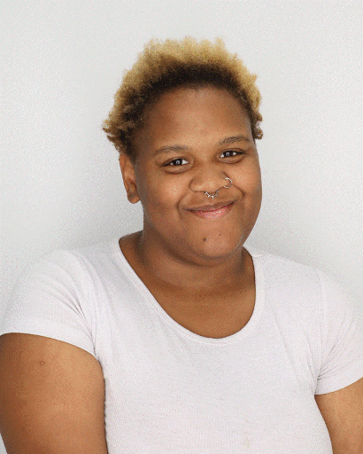 Photograph of a person with short, curly blonde hair wearing a plain white short-sleeve shirt against a white background. The person's face is obscured by a blur, focusing attention on hairstyle and clothing.
