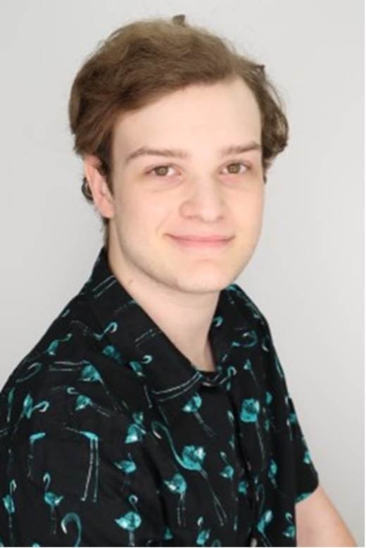 Photograph of a person with medium-length light brown hair wearing a black shirt with a teal flamingo pattern. The person is posed against a plain light gray background with their face blurred for privacy.