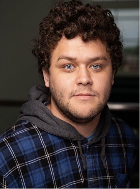Photograph of a person wearing a blue and black plaid shirt layered over a gray hoodie, set against a dark, blurred background. The person's face is obscured by a blur effect, focusing attention on clothing and hair texture.