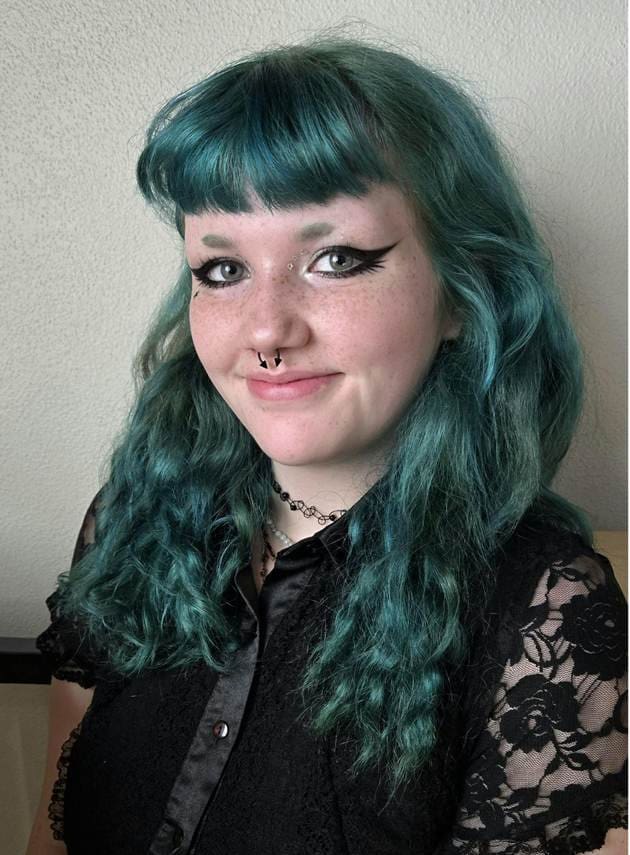 Photograph of a person with long, wavy teal hair wearing a black lace top and a chain necklace. Background features a plain light-colored wall, emphasizing the hairstyle and clothing details.