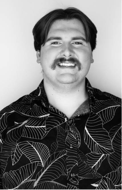 Photograph of a person wearing a black shirt with large white leaf patterns, set against a plain white background. The face is obscured by a gray square, focusing attention on the shirt's design and overall attire.