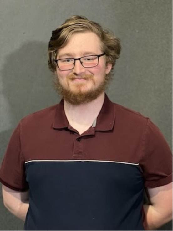 Photograph of a person standing against a plain gray background wearing a short-sleeve polo shirt with maroon upper and navy blue lower sections separated by a thin white stripe. The person's face is blurred, and their hands are positioned behind their back.