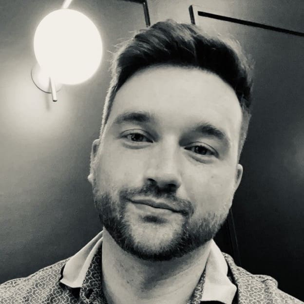 Photograph of a person sitting indoors with a round wall light fixture glowing brightly behind their head, creating a halo-like effect. The background features dark walls with framed artwork, and the person wears a patterned shirt with a collar.