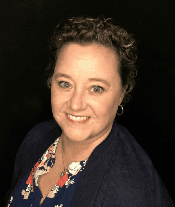 Photograph of a person with curly hair wearing a floral-patterned shirt and dark cardigan against a black background. The face is blurred to obscure identity, focusing on clothing and hairstyle details
