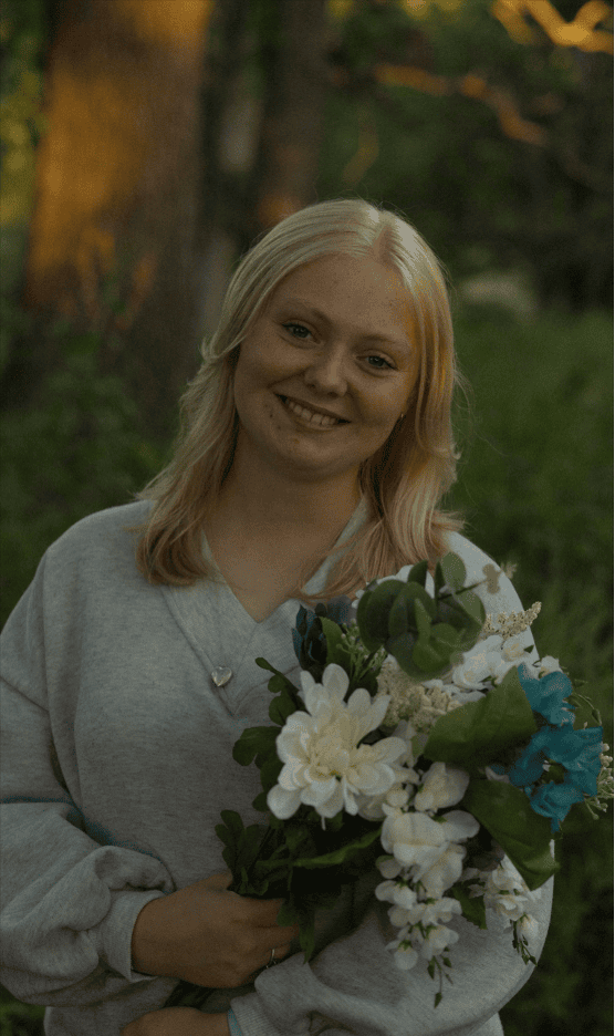 Photograph of a person with blonde hair wearing a light gray sweatshirt holding a bouquet of white and blue flowers with green leaves. Background features blurred trees and greenery, suggesting an outdoor setting during golden hour