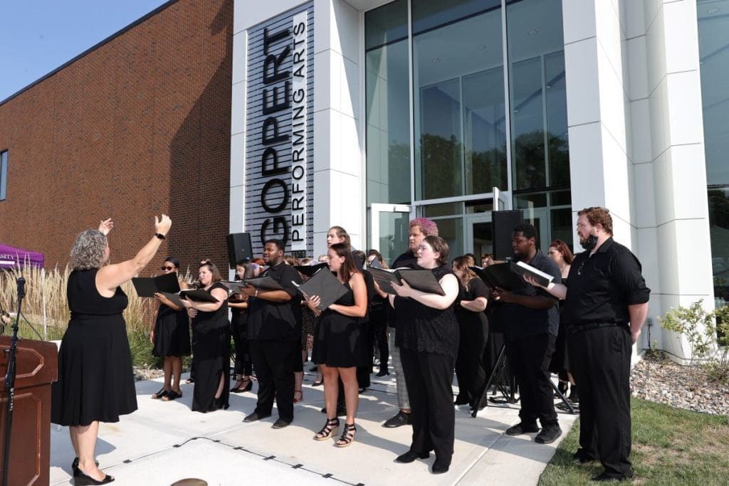 A choir of approximately 20 students dressed in black performs outdoors in front of the Goppert Performing Arts Center at Avila University on a sunny day. A conductor stands at left with her back to the camera, arms raised, leading the group. Students hold black folders and sing together near the building's modern glass entrance. A podium with a microphone is visible to the left.