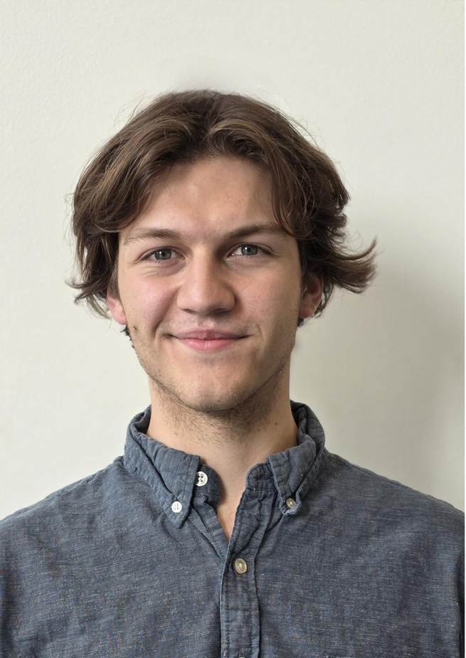 Photograph of a person wearing a blue button-up shirt standing against a plain light-colored background. The person's face is blurred, highlighting the casual attire and hairstyle.