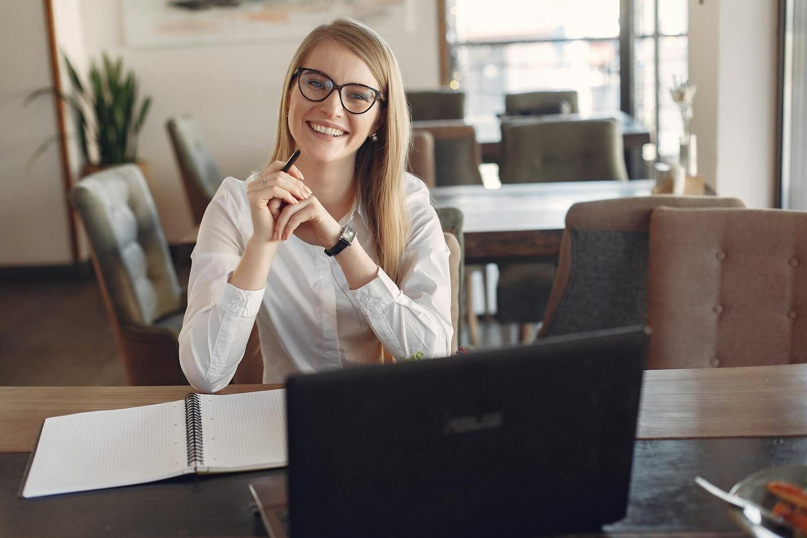 Young businesswoman wearing glasses works at a café with a laptop and notebook.