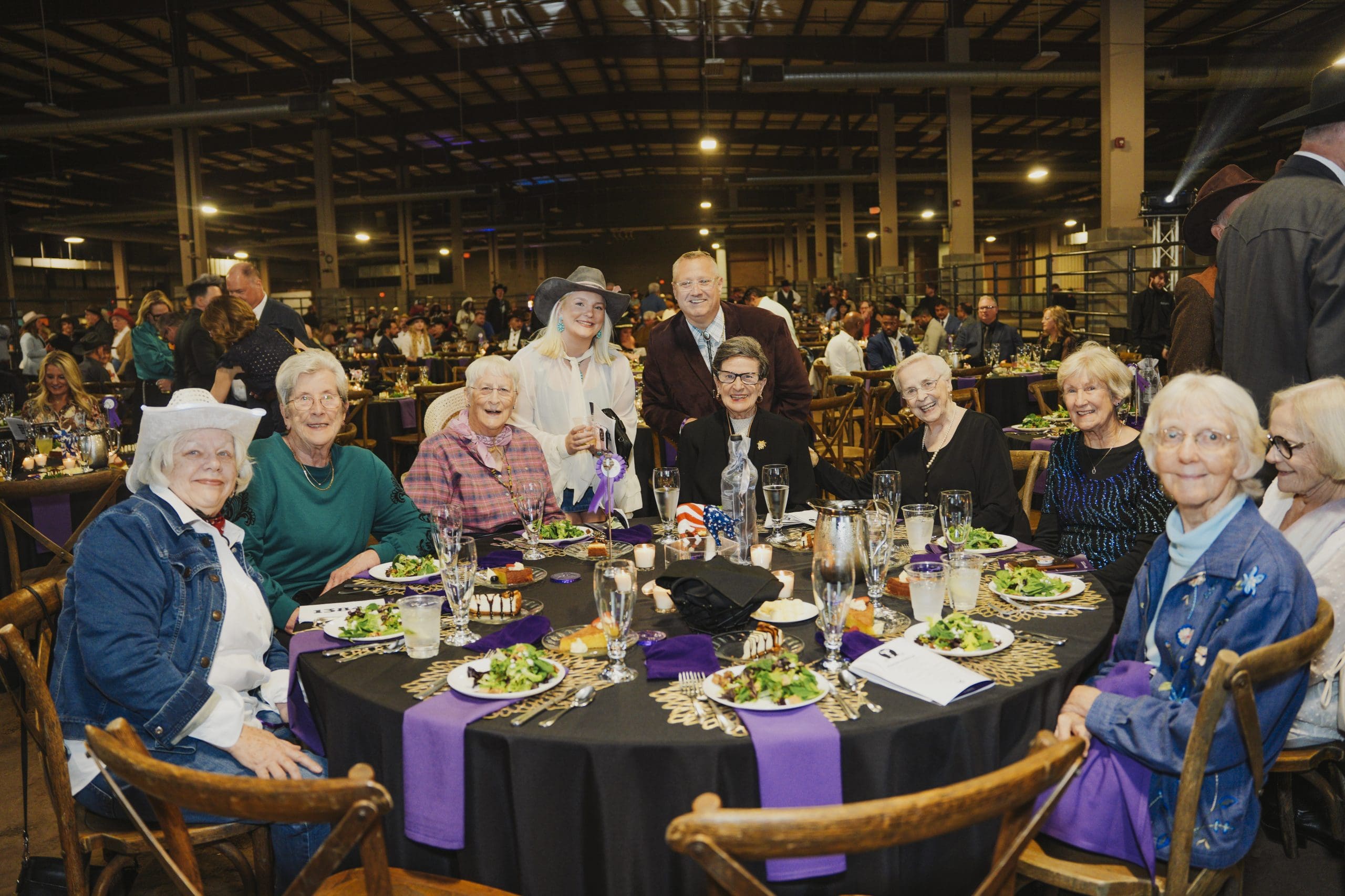 A group of Avila University supporters and guests smile together at a round dinner table elegantly set with candles, floral centerpieces, and purple linens at the 50th Steer Dinner gala.