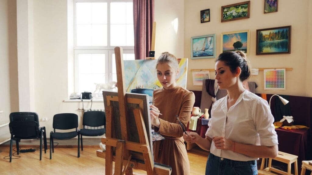 Two women painting on canvas in a well-lit art studio filled with paintings on walls.