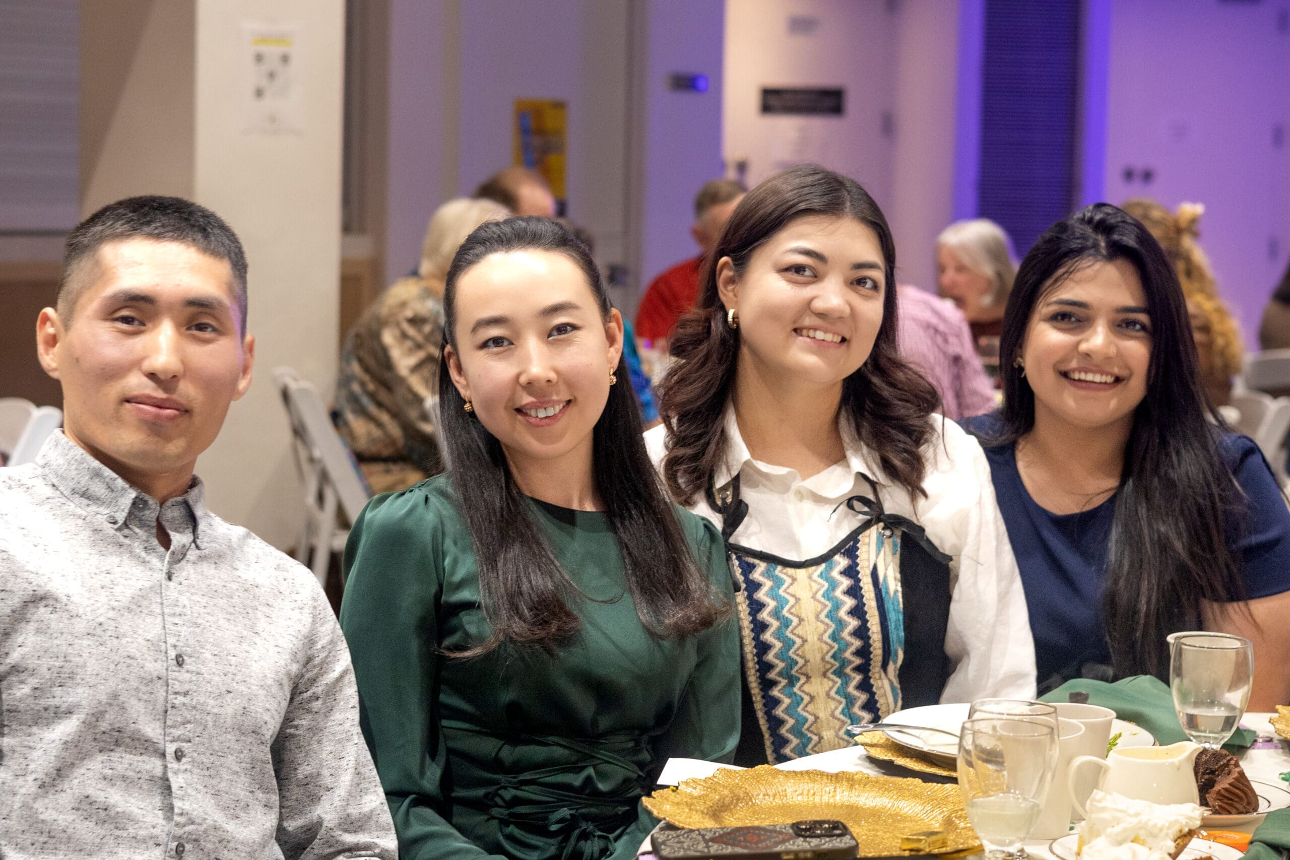 Guests smile at a table set with gold chargers and desserts during Avila Dinner Theatre, with other attendees dining under purple lighting behind them.